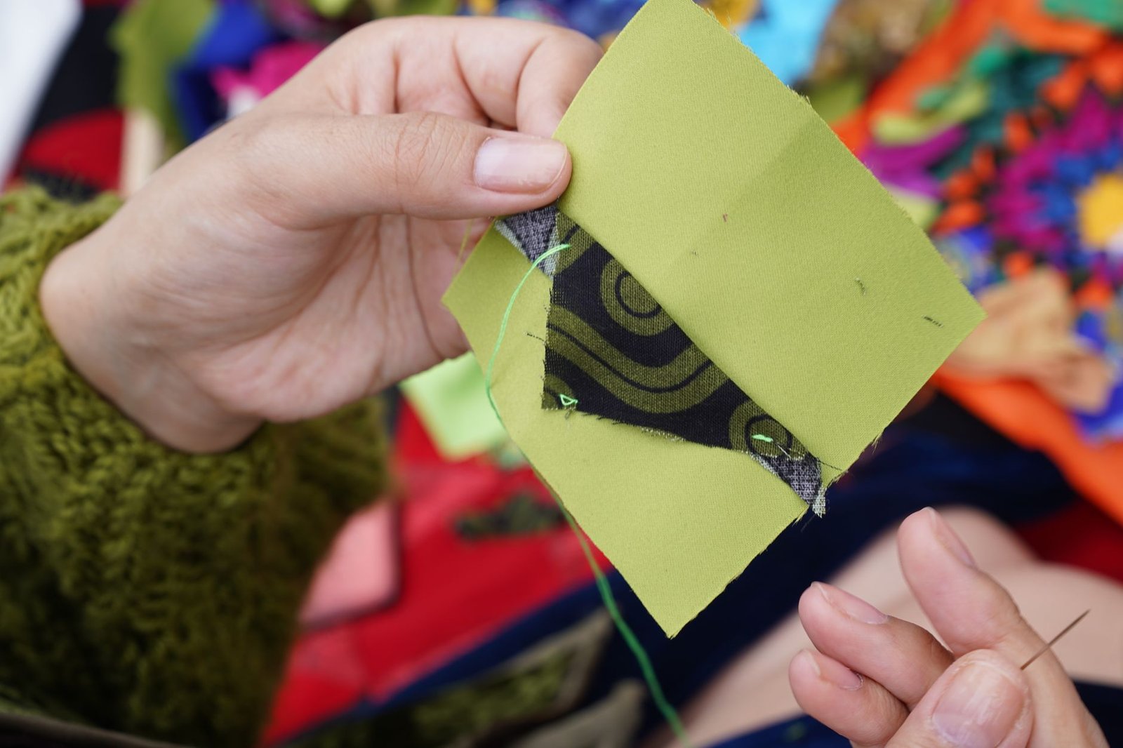 Hands piecing a patterned green fabric square with needle and thread