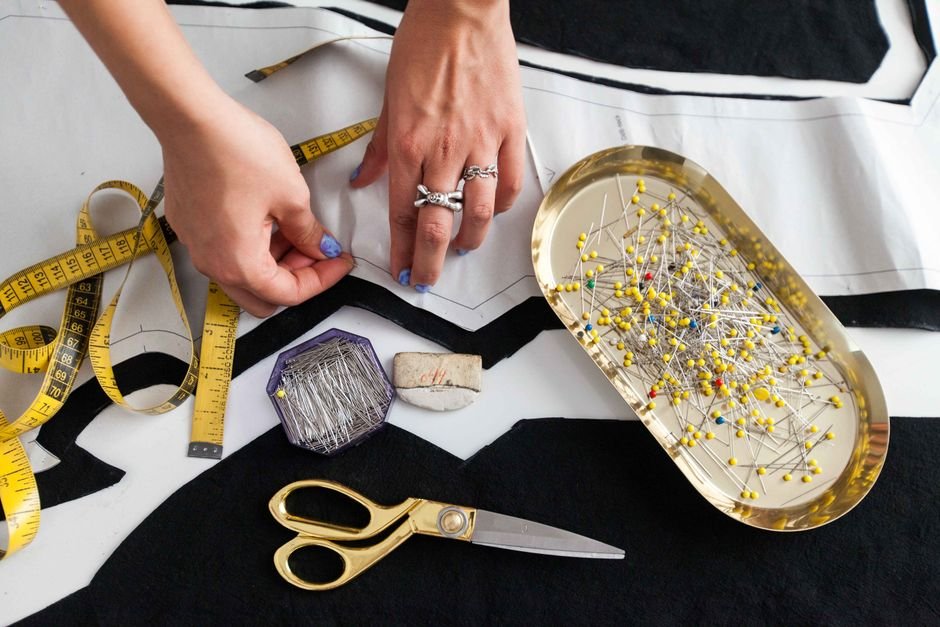 Hands positioning fabric pattern pieces beside a tray of sewing pins and scissors